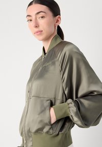 Olivengrønn bomberjakke i glatt sateng, med glidelås, ribbede mansjetter og sidelommer. "Alaïa"-logo brodert på brystet.