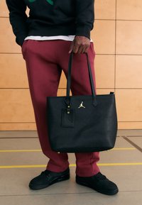 Svart toteveske med teksturert mønster, gullfarget metall, og to håndtak. Kombinert med mørkerøde joggebukser og svarte joggesko.