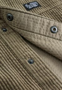 Chemise en coton vert olive avec tissu côtelé texturé. Dotée de boutons-pression et d'une étiquette sur le col. Doublure intérieure lisse.