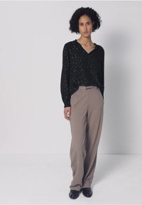 Schwarze Langarmbluse mit weißen Sprenkeln, kombiniert mit hoch taillierten beigen Hosen. Mit entspanntem Schnitt und glattem Stoff. Schwarze Schuhe.