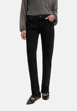 Jean en denim noir avec une coupe droite, des détails légèrement déchirés et un design à cinq poches. Associé à un haut en tricot gris et des ballerines noires à bout pointu.