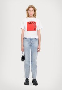 Hvit bomullst-skjorte med rødt logo-grafikk, kombinert med lyseblå denimbukser og svarte klumpete sko, holder en liten svart veske.