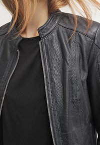 Svart läderjacka med strukturerade paneler och silverdragkedja, med rund halsringning och en tight passform. Buren över en svart topp.