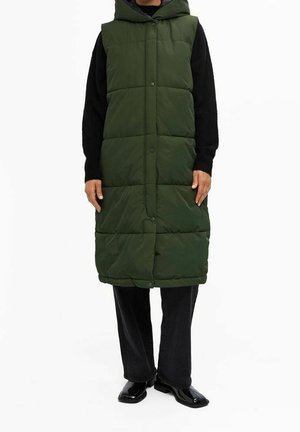 Osoba nosící dlouhou tmavě zelenou prošívanou vestu bez rukávů s kapucí přes černé tričko s dlouhým rukávem, černé kalhoty a černé boty.