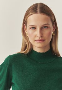 Zielony, błyszczący sweter z dzianiny o wysokim kołnierzu i fakturowanej tkaninie. Złote kolczyki dodają kontrastującego metalicznego akcentu. Gładkie, jasne tło.