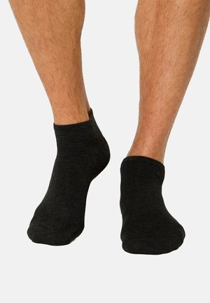 Kaksi paljasta jalkaa, joissa mustat nilkkasukat, yksinkertaista harmaata taustaa vasten. Näytetään sukien etu- ja sivunäkymät.