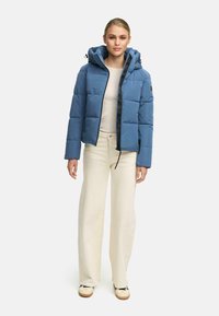 Doudoune bleu clair avec capuche, dotée d'une fermeture éclair à l'avant, de poignets élastiques et d'un design matelassé. Portée sur un haut beige et un pantalon crème.