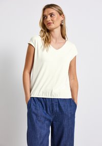 Haut léger en crème à manches courtes avec un décolleté en V et un ourlet froncé, associé à un pantalon en denim bleu ample, formant une tenue décontractée.