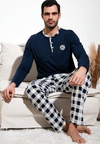 Mornarska majica dugih rukava s gumbima na izrezu i bijelim kariranim hlačama za pijamu. Lagani materijal s teksturiranom bijelom sofom u pozadini.