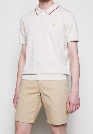 Mand iført beige shorts og en cremefarvet polo med røde kanter på kraven og et lille gult logo på brystet.