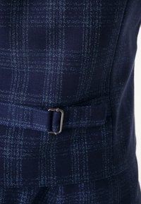 Gros plan d’un tissu bleu marine à motif à carreaux bleu clair et d’une ceinture ajustable avec une boucle rectangulaire argentée.