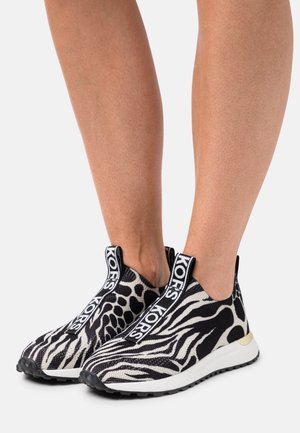 Beine mit schwarz-weißem Animal-Print-Slipper-Sneakern, die auf den vorderen Zuglaschen "KORS"-Markenaufdruck und weiße Gummisohlen haben.