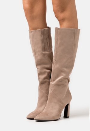 Femme portant des bottes en suede taupe à talons hauteur genou avec fermeture éclair sur le côté et bouts pointus, se tenant sur un fond blanc.