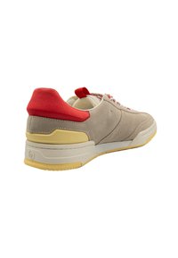 Sneaker beige in suede con tallone in tessuto rosso, intersuola bianca e suola in gomma gialla. Presenta lacci rossi e bianchi e un'etichetta di trazione.