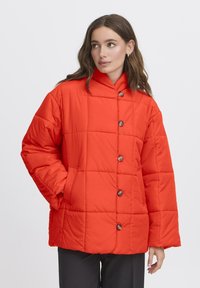 Kvinde med langt brunt hår iført en lys orange quilted jakke med knapper, hænderne i lommerne, ser til siden på en hvid baggrund.