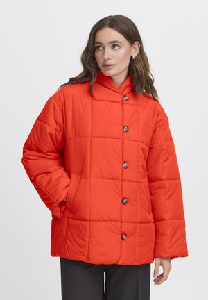 Frau mit langen braunen Haaren, die eine leuchtend orange gesteppten Jacke mit Knöpfen trägt, die Hände in den Taschen, seitlich blickend vor einem weißen Hintergrund.