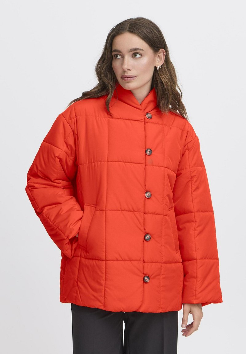 Kvinde med langt brunt hår iført en lys orange quilted jakke med knapper, hænderne i lommerne, ser til siden på en hvid baggrund.