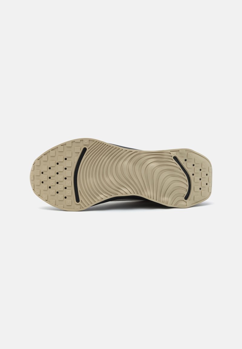 Suela de zapatilla negra con goma texturizada, que presenta un patrón en ondas y secciones de agarre en forma de cuadrado. Contraste de color con acentos beige.