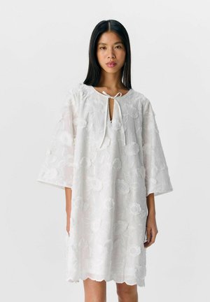 White dress with floral embroidery, short sleeves, keyhole neckline, and a tie detail. The fabric is lightweight and sheer, with a scalloped hem.