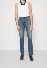 Jeans de mezclilla azul con un acabado desgastado, pierna recta, cinco bolsillos y una bragueta de botones, combinados con una camiseta sin mangas blanca y botas negras hasta el tobillo.