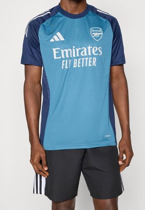 Mand iført blå Arsenal fodboldtrøje med Emirates Fly Better-logo og sorte Adidas shorts med hvide striber, stående mod hvid baggrund.