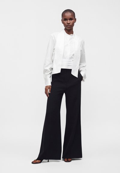 Femme portant un chemisier blanc à manches longues avec un panneau avant et un pantalon noir à jambes larges, debout avec une main dans la poche sur fond blanc.