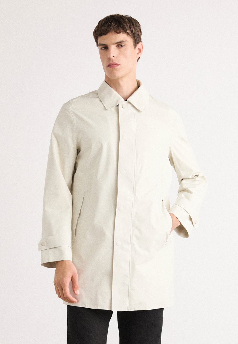 Trench coat de un beige claro con una tapeta de botones, gran cuello y bolsillos con solapa. Presenta un corte recto y puños abotonados. Se lleva sobre pantalones oscuros.