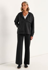 Zwarte zip-up jas met een capuchon, voorzien van een gladde textuur en twee voorzakken, gecombineerd met ruime zwarte broek en zwarte sneakers.