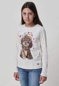 Mädchen mit langen braunen Haaren, das ein weißes Langarmshirt mit einem Pudel in Sonnenbrillen und roten Herzen auf der Vorderseite trägt, sowie eine blaue Jeans.