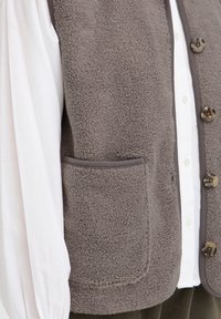Gros plan d'un gilet polaire marron texturé avec des boutons sur une manche de chemise blanche et une poche avant visible.