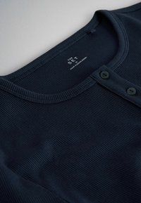 Cămașă albastru marin cu rețea, cu decolteu rotund și doi nasturi negri pe plastronul din față, etichetată "The Set, Made in Bangladesh."