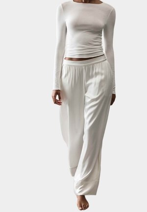 Persona vestida con una camiseta blanca ajustada de manga larga y pantalones anchos de satén blanco, caminando descalza sobre un fondo liso.