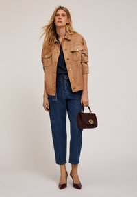 Chaqueta de ante marrón claro con grandes bolsillos, combinada con vaqueros de tiro alto azules. Un bolso burdeos y tacones de punta completan el look.