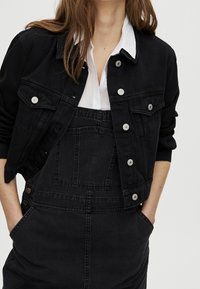 Černá denimová bunda s knoflíkovým zapínáním, náprsními kapsami a límcovým designem, přes černé denimové overaly. Měkká textura a casual fit.