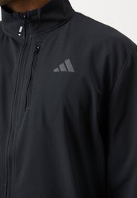 Veste de sport noire avec une poche zippée sur la poitrine et un logo Adidas réfléchissant sur le côté gauche.
