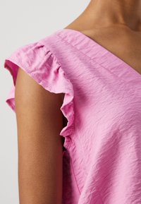 Nära bild av en person som bär en rosa tygblus med volangärm och V-ringning, med ljus hudton.