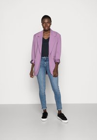 Blazer oversized lavanda com mangas enroladas, combinado com uma camisola preta de decote em V, jeans skinny azul claro e sneakers pretos tipo slip-on.