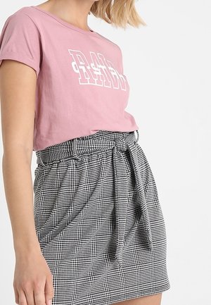 Kobieta ubrana w różowy krótki T-shirt z białym napisem oraz wysoką spódnicę w czarno-białą kratę, zawiązaną materiałowym paskiem.