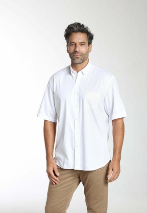 Homme aux cheveux foncés et à la barbe, portant une chemise blanche à manches courtes boutonnée et un pantalon beige, debout devant un fond clair.