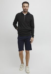 Crna zip-up hoodie s crvenim akcentima, uparena s mornarskim šortsevima. Model nosi bijele tenisice. Glatka tkanina, ležeran kroj, minimalistički dizajn.