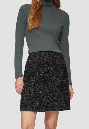 Femme portant un col roulé à manches longues gris foncé et une jupe noire texturée jusqu'aux genoux, sur un fond uni.