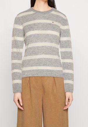 Femme portant un pull rayé horizontal gris et blanc et un pantalon marron ample, debout devant un fond uni.