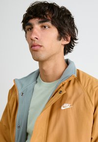 Nike dzseki, amelyen egy nagy, tompa narancssárga külső réteg található, világoskék polár gallérral, fehér logóval, és egy puha zöld póló van alatta.