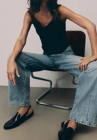 Top de tirantes negro, jeans de cintura alta azul claro y zapatos de tacón negro con correa al talón. Modelo sentado con las piernas abiertas en una silla minimalista.