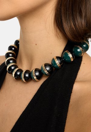 Kobieta nosząca masywny naszyjnik z naprzemiennymi czarnymi i ciemnozielonymi błyszczącymi koralikami oplecionymi złotymi skręconymi opaskami, w połączeniu z czarną bluzką.