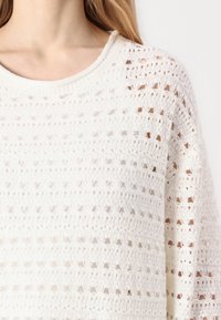 White knitted sweater featuring an open, textured design with a wide neckline and dropped shoulders, showcasing horizontal and vertical patterns.