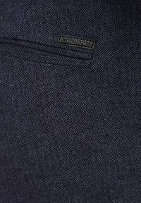 Tamno plavi tkanina u šaru riblje kosti s suptilnom teksturom. Na njemu je pričvršćena crna oznaka s natpisom "JACK & JONES". Ima elegantno dizajniranu džep.