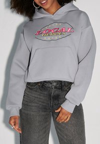 Szara, krótka bluza z kapturem z przednią kieszenią, z wielokolorowym napisem "LOCAL HEROES" oraz akcentami w kształcie gwiazdy. Materiał wydaje się być miękki i casualowy.