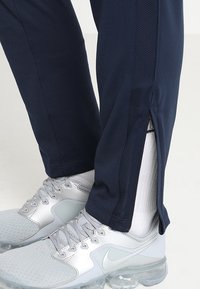 Marina sportbyxor med nätpaneler på sidorna och dragkedja vid nederkant, i kombination med ljusgrå sneakers med genomskinlig sula.