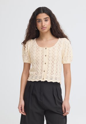 Kvinde med langt krøllet hår iført cremefarvet kortærmet strikbluse med knapper og sorte højtaljede plisserede bukser, stående foran ensfarvet baggrund.